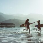Three people on a beach, running into the ocean with surboards in a tropical climate, with mountains in the background.