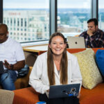 Three people sit on an orange couch and work on laptops.