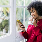 Woman in a red shirt checks her phone inside on a rainy day while holding a white coffee mug / email deliverability