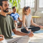 A man with a ponytail meditates in a yoga class, surrounded by other yoga class participants in a bright studio with large windows and plants.